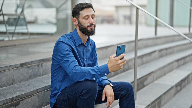Young hispanic man business worker sitting on stairs make selfie by smartphone at street