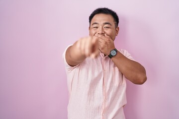 Chinese young man standing over pink background laughing at you, pointing finger to the camera with...