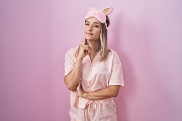 Blonde caucasian woman wearing sleep mask and pajama thinking concentrated about doubt with finger on chin and looking up wondering