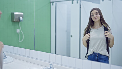 Young beautiful girl student wearing backpack looking on mirror at bathroom
