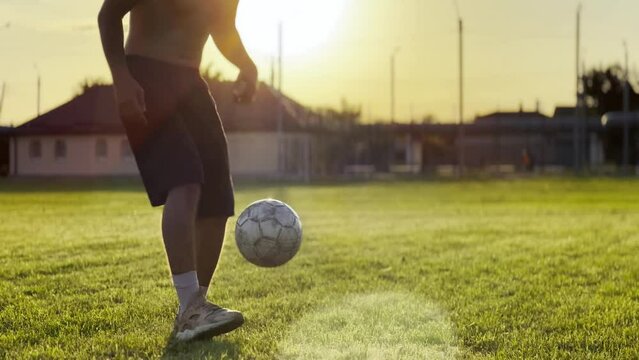 Male feet of professional footballer kicking ball at green field. Legs of young man juggling soccer ball on stadium at sunset. Sportsman practicing tricks outdoor. Freestyle football concept