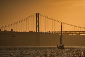 Obraz premium Stunning sunset view of Lisbon, Portugal, with a sailboat sailing across a bridge