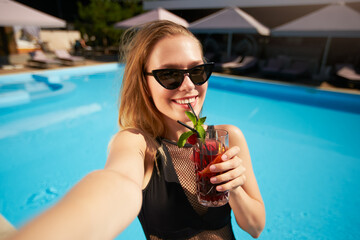 Travel blogger woman in bikini taking selfie photo with cocktail near swimming pool. Pov of...