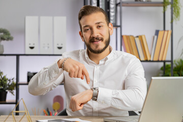 It is your time. Young middle eastern boss business man showing time on wrist watch clock, approve, pointing finger, time to start work, deadline. Freelancer guy at office workplace looking at camera