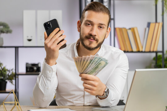 Business man showing smartphone sincerely rejoicing win, receiving money dollar cash banknotes, success lottery luck, recommends earning by phone. Freelancer guy at office workplace looking at camera