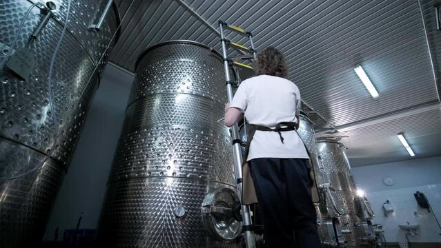 A happy successful farmer or winemaker is walking in the middle of wine tanks and checking a fermentation grade of high quality wine production in a modern winemaking facility or winery.