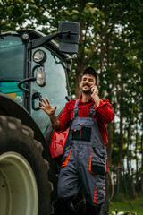 Smiling farmer talking on a mobile phone in front of a tractor in the field. Smart farming concept