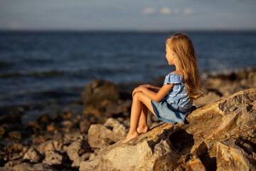 Beautiful girl with long blond hair sits on a stone on the banks of a river, lake, sea.