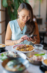 A young Asian woman sits at a table in a cafe and chats with someone on a smartphone.