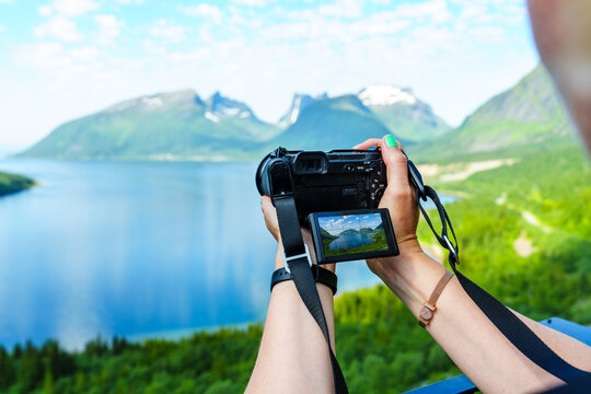 Young woman photographer making landscape photograpy of Norway fjords in bright summer day. Young woman with standing on a hill with green grass and making snapshot of a beautiful scene, arial view