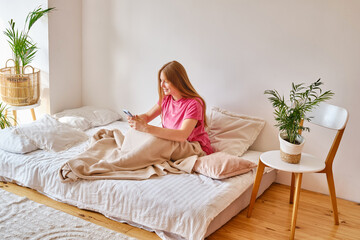 Girl holds a smartphone in her hands in a comfortable bed and checks the social network account before going to bed or after waking up. Gadget addiction. Woman sending text message on mobile phone