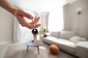 Young female hand holding keys against new bright interior with sofa. housewarming concept
