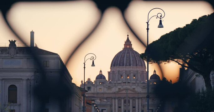 Amazing panoramic cinematic view through the bridge fence of St. Peter's Basilica, Vatican City in Rome, Italy at vibrant orange sunset sky