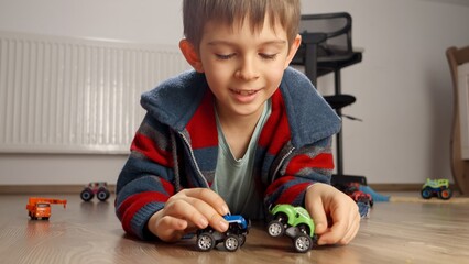 Portrait of cute boy lying on floor and playing with two toy cars. Children playing alone, development and education, games at home.