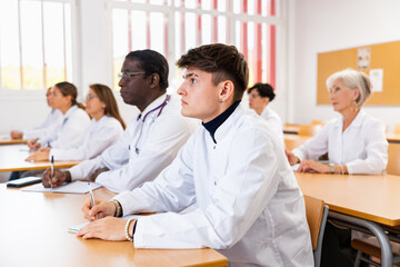 Fototapeta na wymiar Young man in white coat sitting at desk in classroom, attending lecture in medical university with group of students and colleagues.