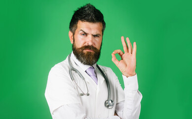 Surgeon doctor showing ok sign with fingers. Bearded physician man with stethoscope in white coat gesturing excellent. Serious confident male doctor in medical uniform showing okay, approval gesture.