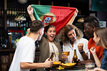 Excited diverse soccer supporters with flag of Portugal watching tournament with pint of beer and chips in the pub
