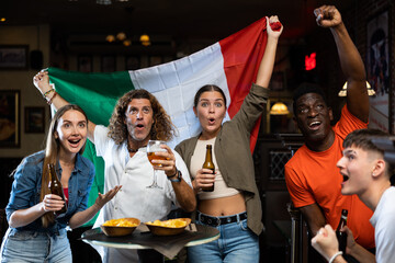 Fototapeta premium Happy multiracial friends holding the flag of Italy, rejoicing winning football game and drinking beer in the bar