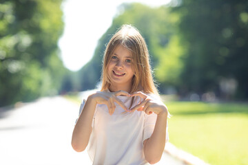 Kindness Concept. Portrait of cute blonde hair girl making heart shape with her hands and fingers near chest, smiling to camera, posing while walking outdoor in a park, copy space
