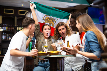 Excited young adults of different nationalities, sports fans celebrating victory of favorite team with beer in bar, waving national flag of Iran
