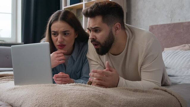 Caucasian family couple upset disappointed read bad news negative result refusal bank email upset with failure shocked woman and man homeowners on bed use laptop computer watch scary movie in bedroom