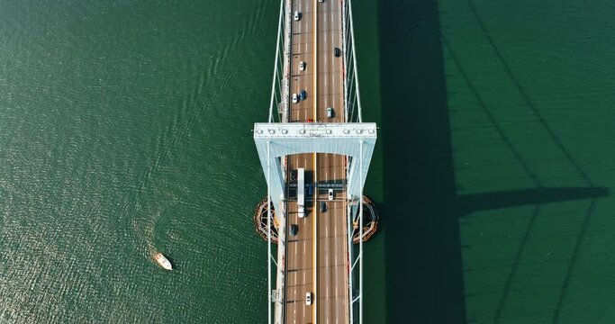 Rising above the support of suspension bridge. Numerous cars run quickly by the roads on the bridge. Yacht sails by the green water. Top view.
