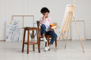 Young woman holding brush and artist`s palette near easel with canvas