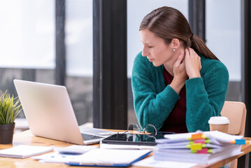 Young businesswoman office worker suffering from neck pain, massaging her neck at office table workplace, feeling overworked.