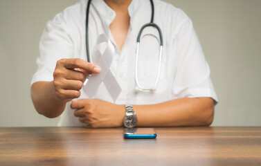 Physician holding a white ribbon symbol the fight against lung cancer while sitting at the table in the hospital