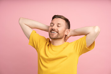 Relaxed man holding hands behind head smiling with closed eyes standing on pink background....
