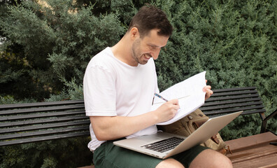 Fototapeta premium Young handsome caucasian man wearing summer casual outfit sitting on bench at public park, using computer laptop, taking notes at notepad.