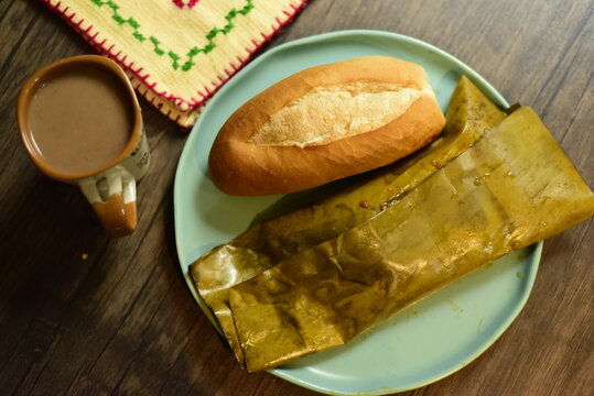 Desayuno de tamal oaxaque&ntilde;o. Con bolillo y atole de chocolate. Plano cenital. Plato azul y una servilleta de tela. Sobre mesa de madera.