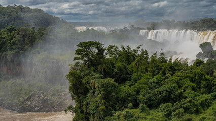 The waterfall flows collapse from the edge of the plateau. Spray, fog. Lush tropical vegetation all around. Clouds in the blue sky. Iguazu Falls. Argentina.