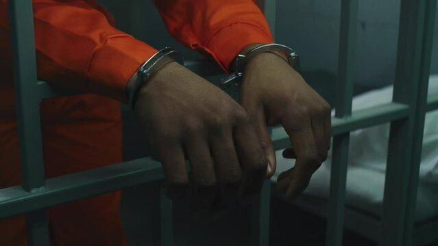 Close up shot of hands in handcuffs leaning on prison cell bars. Criminal serves imprisonment term in correctional facility or detention center. African American prisoner in orange uniform in jail.