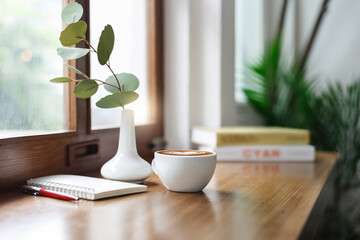 Close-up view, white coffee cup on wooden counter and note book, pen with ceramic vase and small...