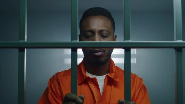 African American man in orange uniform keeps hands in handcuffs on jail cell bars and looks at camera. Sad criminal serves imprisonment term in prison. Prisoner in correctional facility. Dolly shot.
