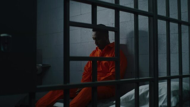 Guilty African American man in orange uniform sits on prison bed and thinks about freedom. Gloomy criminal in correctional facility or detention center. Prisoner serves imprisonment term in jail cell.