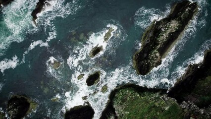 Aerial top down angle side dolly drone footage of ocean blue waves break on high cliff of a rocky mountain. Bow fiddle rock, Scotland