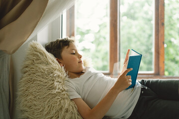 Cute boy reading a book on the windowsill near the window. Child relaxing at home on sunny day...