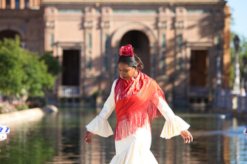 Obraz premium Young black woman dressed as a flamenco gypsy in a famous square in Seville, Spain. She is wearing a beige dress with ruffles and a red shawl and is in front of a canal in the square.