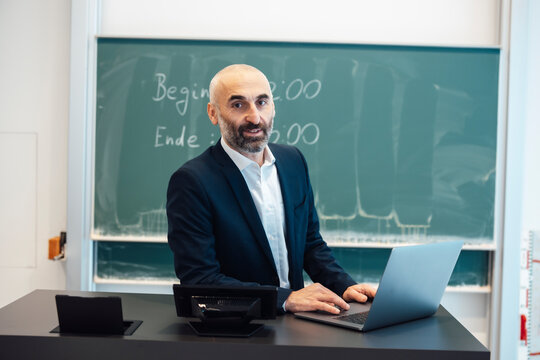 Professor sitting at desk with laptop in lecture hall