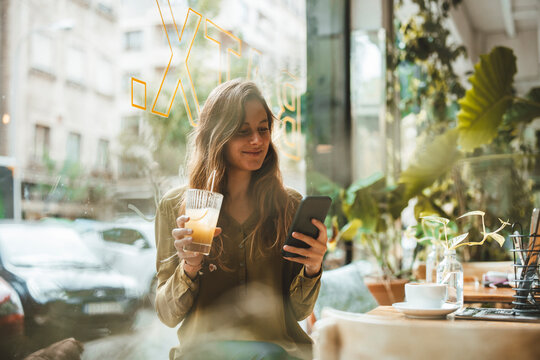 Smiling young woman with juice using smart phone in cafe