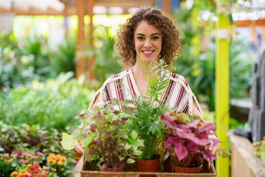 Woman with curly hair holding potted plants in tray at nursery