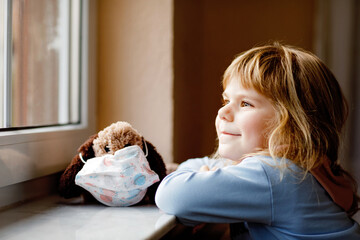 Cute toddler girl sitting by window and looking out. Dreaming child put a medical mask on soft dog...