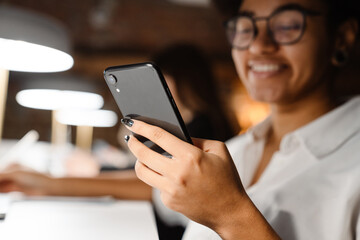 Smiling african woman using smartphone while working on laptop in office