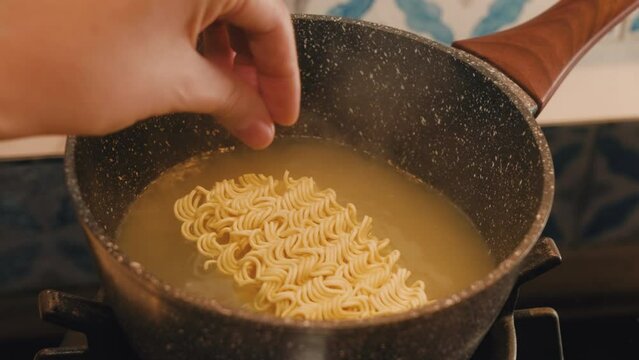 POV hands cooking instant noodle soup at home. Female hand putting dry noodles into boiling water in a pan at home and stirring. Delicious and simple snack. Close-up. Steaming food on gas stove.