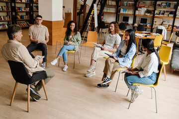 Adult man lecturer communicating with group of students while sitting in library