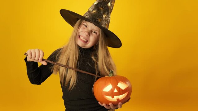 Little girl in witch hat with magic wand isolated on yellow background. Cute witch carrying orange pumpkin lantern. Halloween holiday. Female laughing with scary pumpkin in hands. 4K, UHD