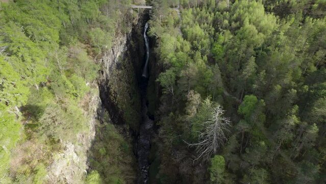 Aerial drone shot flying away from  waterfall inside narrow Corrieshalloch gorge surrounded by forest in Scottish Highlands