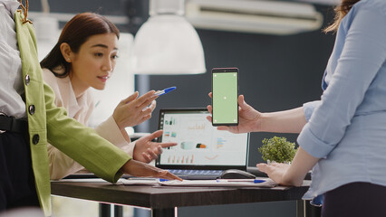 Diverse group of employees working with greenscreen on smartphone, using isolated display with chroma key. Startup workers checking mockup template in business coworking space. Handheld shot.